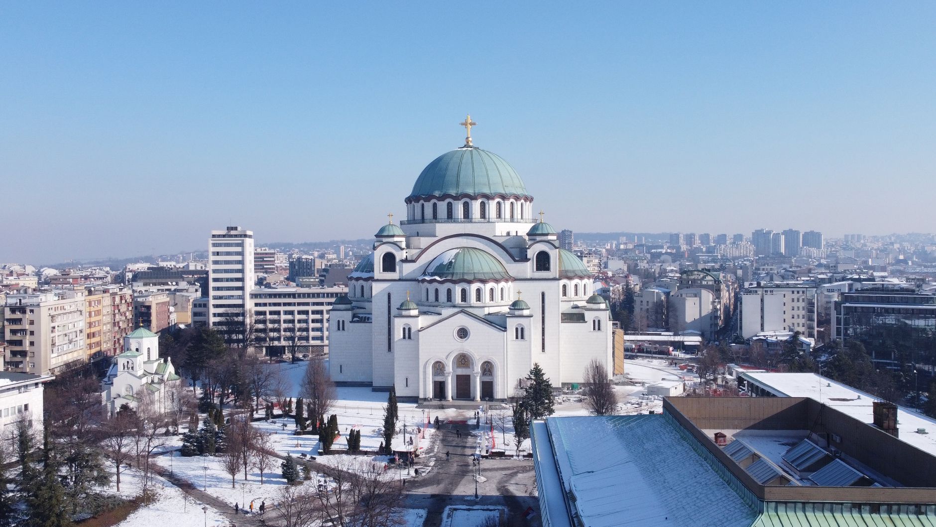 Храм Святого Саввы в Белграде зимой: белый собор с зелёными куполами, заснеженный парк вокруг и город в морозной дымке
